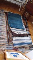 Rugs, mats, and pillow cover stacked on top of a large striped woven rug on wooden floor, showing texture, colors, and tassels.
