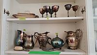 Silver-plated pitchers, teapot, dressser brush, and vegetable dishes on cabinet shelves showing tarnish and wear.