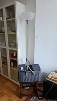 Three-item lot photo showing tall Ikea floor lamp with white glass shade, a wooden stool marked Leslie, and grey wicker basket sitting on stool