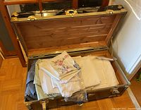 Photo of open metal trunk showing table linens inside, including folded embroidered linen pieces and plain white linens