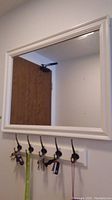 Rectangular white framed mirror with five metal coat hooks holding keys, seen mounted on a wall with a door reflection.