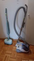 Photo showing Kenmore canister vacuum cleaner with attached hose and telescopic metal wand next to the Eureka floor polisher against a white wall on parquet floor.