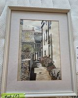 Framed watercolour painting showing British town alley, viewed front with off-white mat and beige wooden frame.