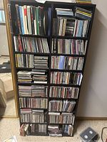 Photo of a black wooden shelf unit packed with many audio CDs in jewel cases, showing multiple compartments filled with CDs.