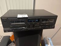 Front view of a black Technics double cassette player unit with two cassette decks, digital display, controls and power button.