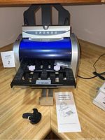 Front angle of folding machine with blue cover, showing fold plates, knobs, and operator manual on countertop alongside an accessory component.