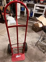 Front view of a red metal moving dolly with curved handle and two black wheels on a concrete floor in a garage setting. Background includes boxes, stools, and shelves with various stored items.