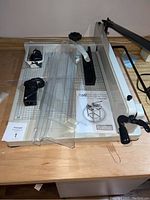 Top angled view of the paper cutter showing base with measurement grid, plastic components, power adapter, and instruction manual beneath plastic pieces.