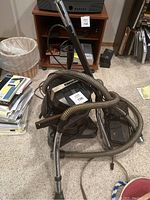 Photo showing Rainbow vacuum cleaner canister with hose coiled around it, metal wand standing upright, and power nozzle attached. Located in basement bedroom with surrounding items.