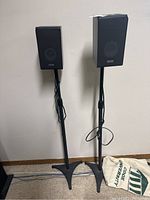 Two Samsung black speakers each mounted on a black adjustable height stand, positioned upright against a wall.