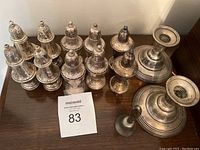Photo showing 13 silver salt and pepper shakers, the bell, and 2 candle holders on a wooden surface with auction lot number card.