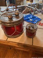Photo showing two vintage drum-shaped containers with red translucent sides and metallic lids topped with drumstick designs, placed on a kitchen counter.