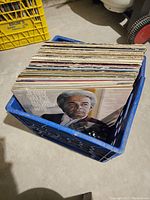 Blue plastic milk crate filled with over 40 vinyl records shown vertically, one front cover visible with a portrait of a man with white hair.