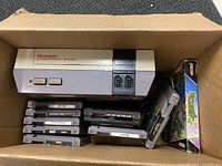 Box containing the Nintendo Entertainment System console and a collection of NES game cartridges stacked inside, some cartridges partially visible, Teenage Mutant Ninja Turtles game box included