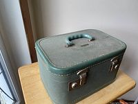 Front and top view of textured green vintage travel case showing handle and metal clasps.