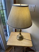 Front view of the vintage lamp on a wooden side table near a window, showing the brass base and beige shade.