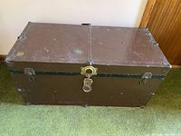 Front view of brown metal vintage trunk chest on green carpet, showing latch and padlock on front.