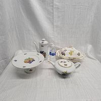 Front view of four antique china pieces including pedestal plate, coffee pot, teapot, and lidded dish. White porcelain with fruit and flower motifs and gold trim.