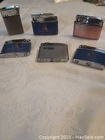 Six assorted vintage lighters on a white cloth surface, including multiple Belvedere branded lighters and one pink textured metal lighter