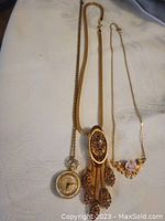 Photo showing three vintage necklaces arranged on white fabric: pendant watch necklace, gold-tone decorative pendant necklace, and porcelain rose accent necklace.