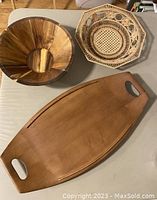 View of three wood items: a large cutting board, a wooden bowl with segmented wood grain, and a woven bowl with open circular designs.