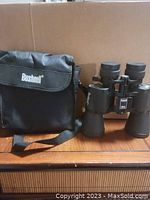 Binoculars standing upright beside the carrying case, all items sitting on a wooden surface with a cardboard background.