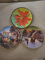 Three decorative plates displayed on a fabric surface: red poinsettia flower plate, children on tree stumps plate, and winter sleigh scene plate.