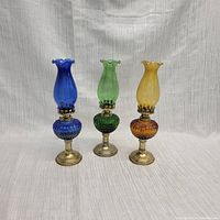 Front view of three hurricane lamps in cobalt blue, emerald green, and amber colors against a neutral background.