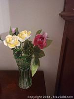 Full view of cut green tinted crystal vase filled with porcelain roses in pink, yellow, and red colors with green leaves on wooden surface.