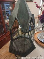 Full view of large metal birdcage with hexagonal base and pointed roof, showing the patina finish and cage bars.
