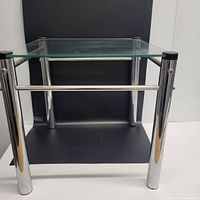 Front angled view of the side table showing the chrome tubular legs, clear glass top, and lower black glass shelf against a black background.
