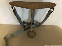 Full view of vintage milking stand/harvest stool showing wooden seat, metal post, footrest base, and leather straps.