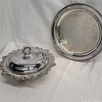 Two silver plated serving pieces including a lidded platter with ornate edges and three legs, and a serving plate with engraved pattern and rope rim.