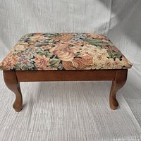 Front-facing view of the small foot stool showing floral tapestry cushion and carved wood legs.