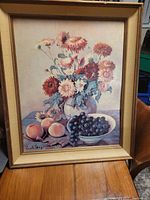 Full view of vintage reproduction still life painting with flowers in vase, peaches, and bowl of grapes within a worn gold frame.
