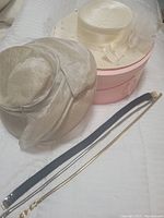 Photo showing two cream-colored summer hats stacked with the pink hat box underneath. Two thin belts placed beside the hats and box.