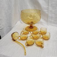 Wide view of amber glass punch bowl with eight matching cups and ladle on white textured fabric background.