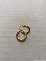 Photo of two small yellow gold hoop earrings with etched pattern, hinged closure and wire latch, arranged on a flat surface