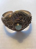 Front angled view of wide silver cuff bracelet showing detailed filigree work and round blue stone in center.