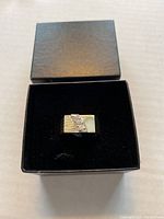 Men's silver-tone ring in a black jewelry box, showing top design with diagonal groove and small stones.
