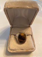 Men's silver ring with large oval tiger's eye gemstone displayed in beige cushioned ring box