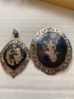 Front view of the silver pin and pendant set with gold-tone designs on black background, showing intricate frames and figures