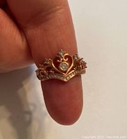 Photo of rose gold tone sterling silver crown motif ring held between fingers showing front view with sparkling stones and heart design.