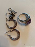 Photo of two pairs of silver earrings and a silver ring with a purple teardrop stone on a flat surface.