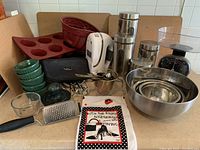 Photo displaying assortment of baking tools including mixing bowls, hand mixer, kitchen scale, canisters, silicon molds, ramekins, measuring spoons, hand grater, muller, and kitchen towel.