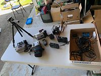 Top view of table with JVC camcorder, tripod, Kodak camera, protective camera cases, remote control, and miscellaneous cables in box.