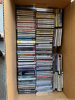 Top view of a cardboard box filled with various music CDs stacked vertically and horizontally, showing a wide range of genres and artists.