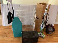 Photo shows the full lot including two laundry hampers (one wooden rectangular and one white wire), ironing board, metal wire organizer, sewing basket, BISSELL floor and spot cleaners.