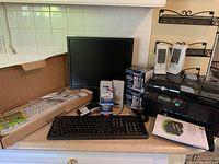 Photo showing a black computer monitor, HP printer, keyboards including white Logitech keyboard, black mouse, power cables, and ink cartridges arranged on a white surface.