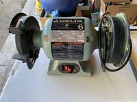 Front view of Delta 6-inch bench grinder showing grinding wheel, wire wheel, safety guard, tool rest, and power switch.
